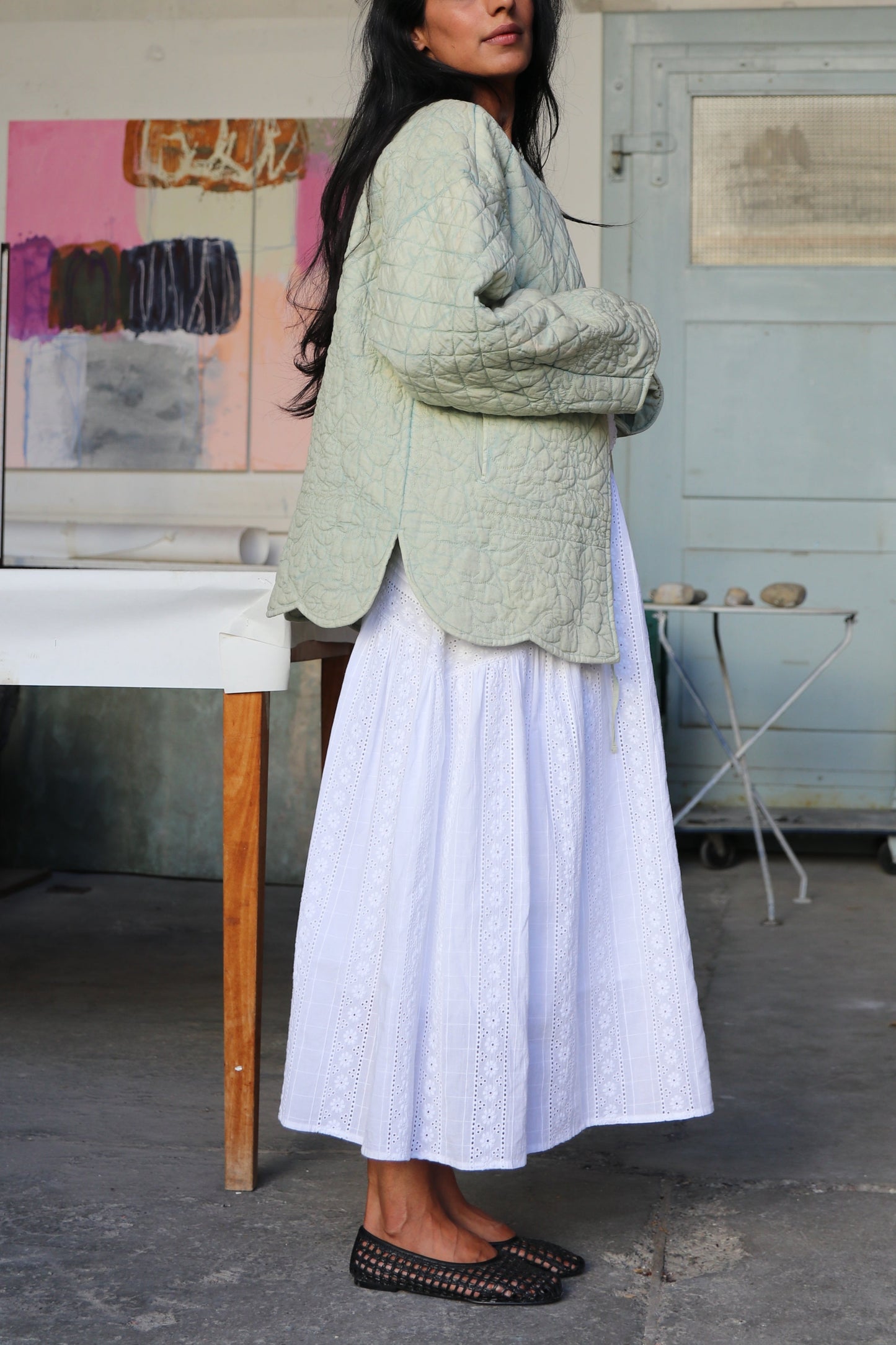 Woman wearing a green quilted jacket and white skirt standing in an artist's workshop