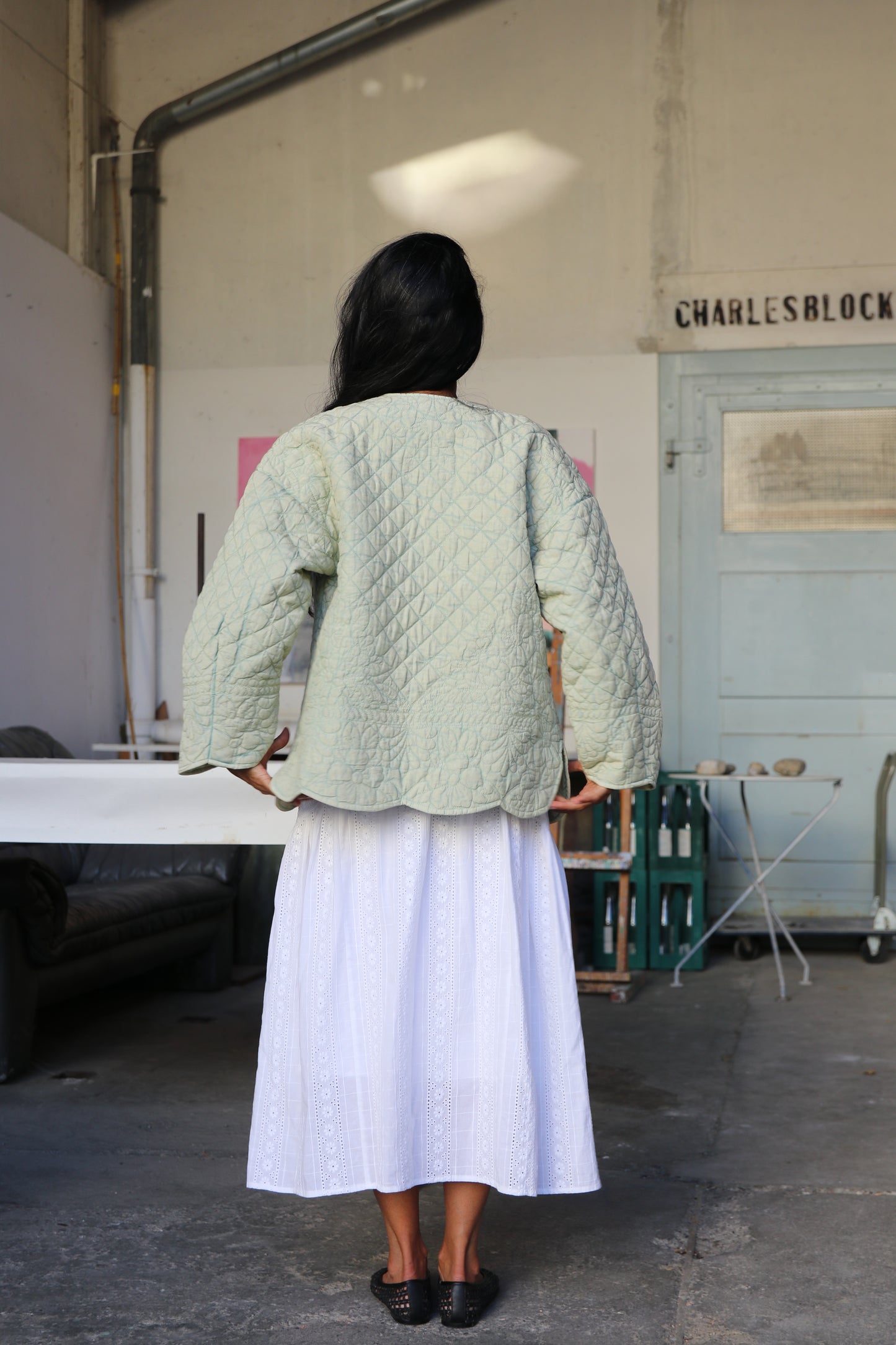 Woman wearing a green quilted jacket and white skirt standing in an artist's workshop.