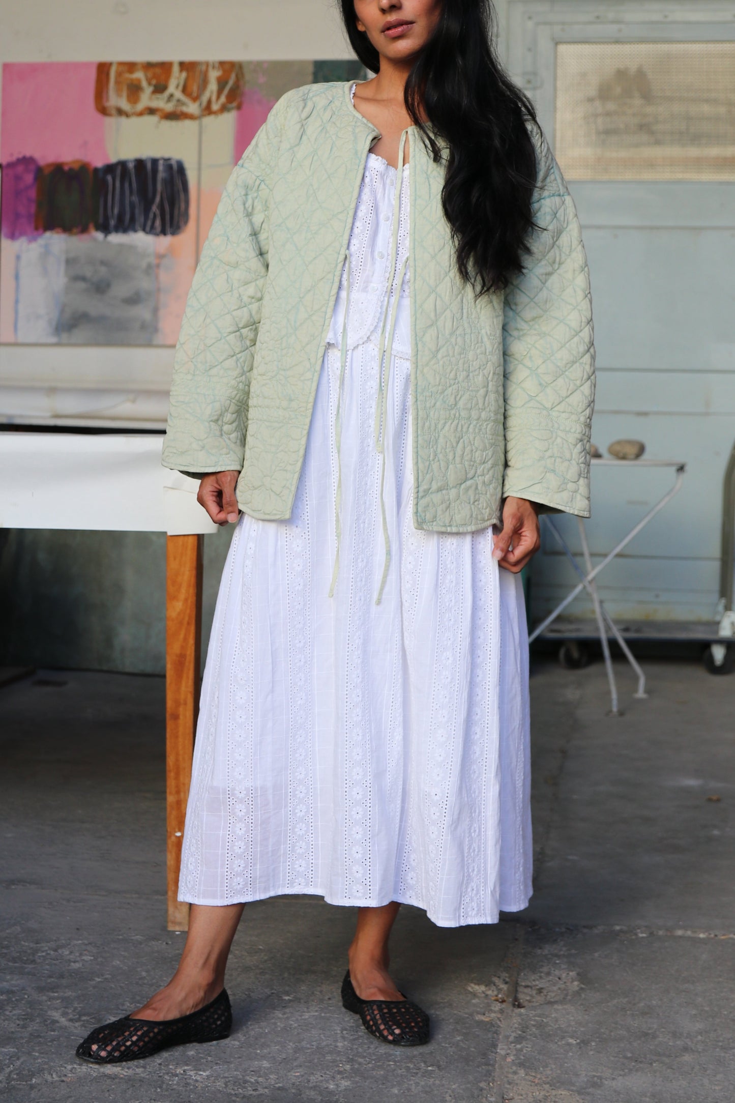 Woman wearing a green quilted jacket and white skirt standing in an artist's workshop.