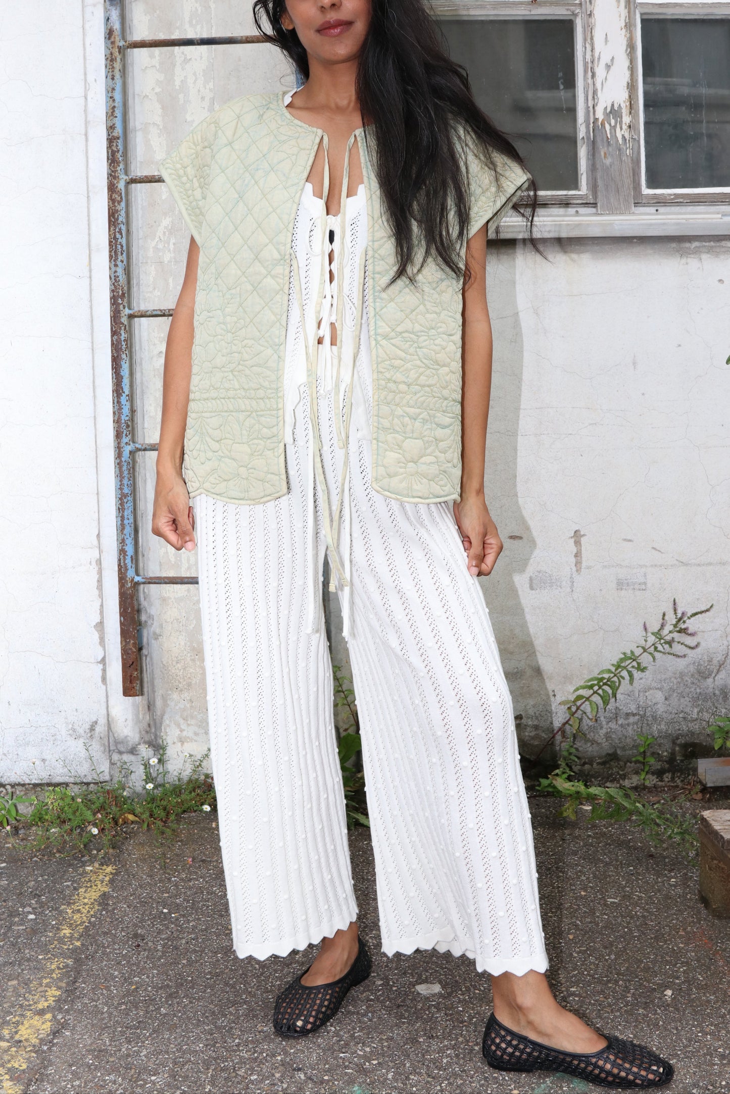 Woman wearing a light green vest with white knit pants standing in front of a building