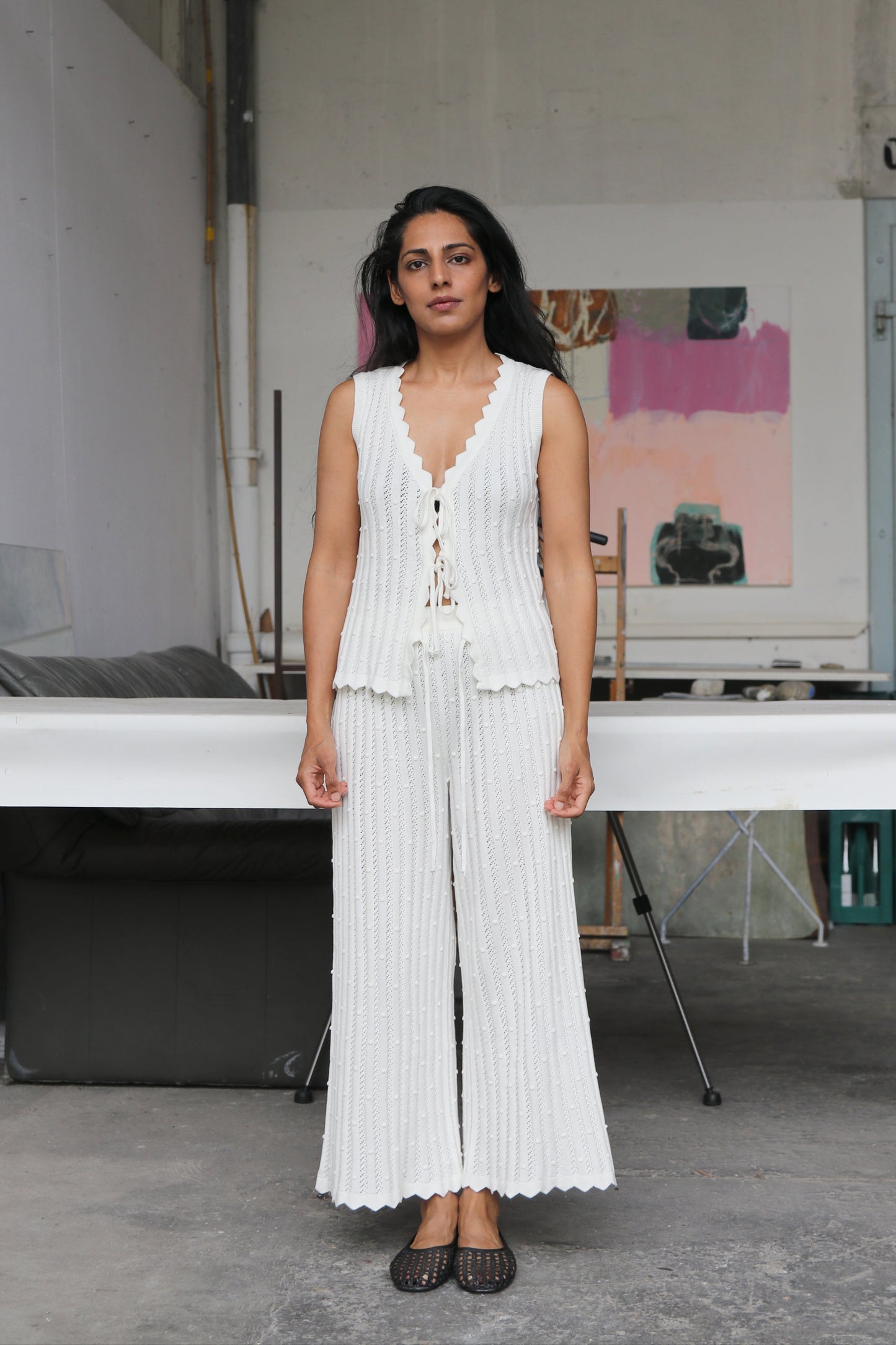 Woman in a white outfit standing in an indoor setting with a white table and wall art in the background.