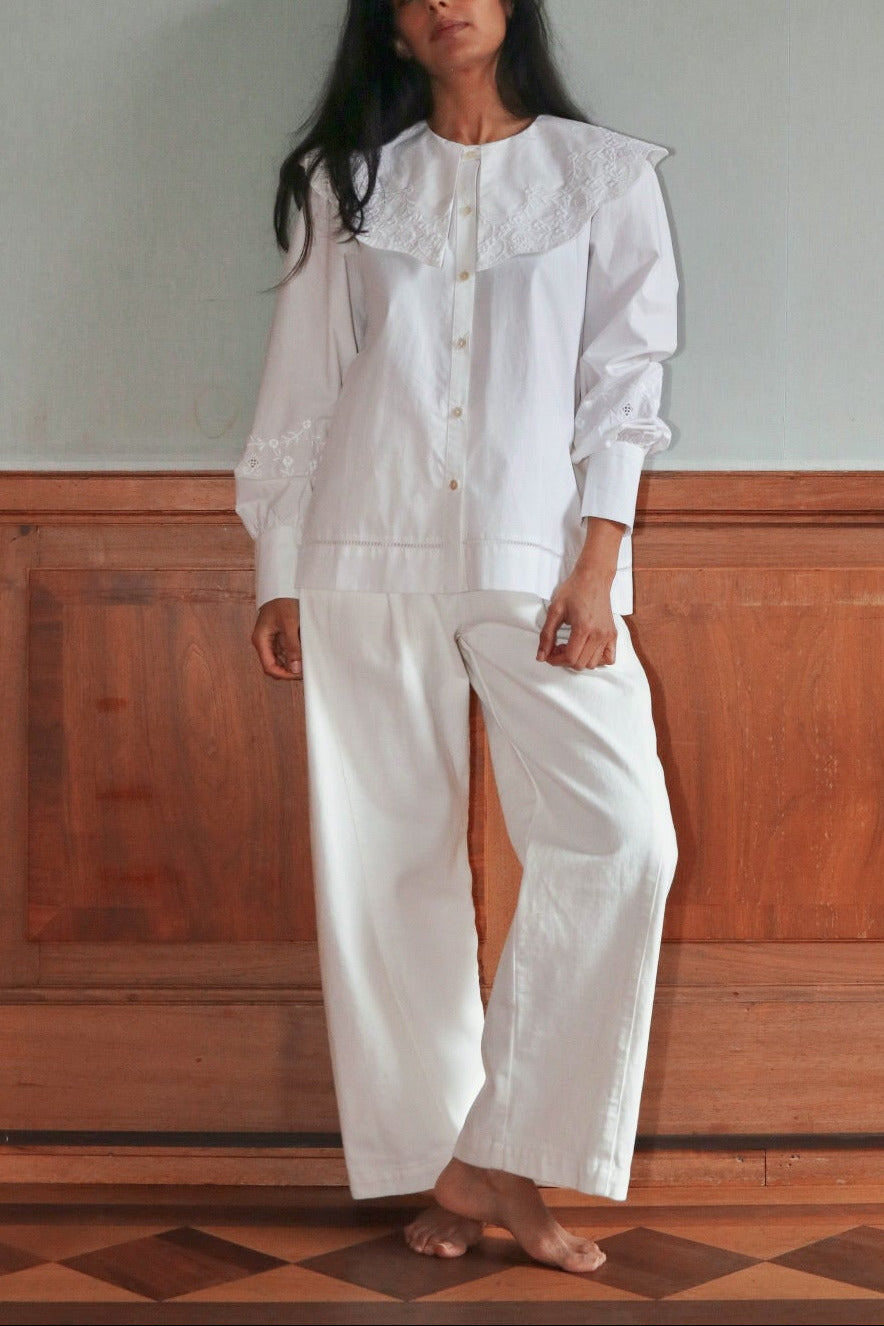 Woman wearing a white embroidered shirt and trouser outfit standing against a plain wall with wooden paneling.