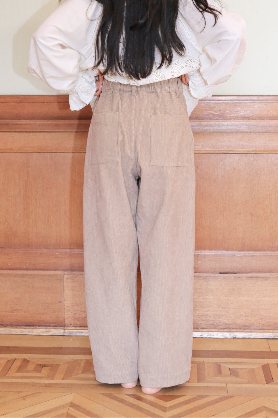 Person wearing a white blouse and beige pants standing in front of a wooden panel.