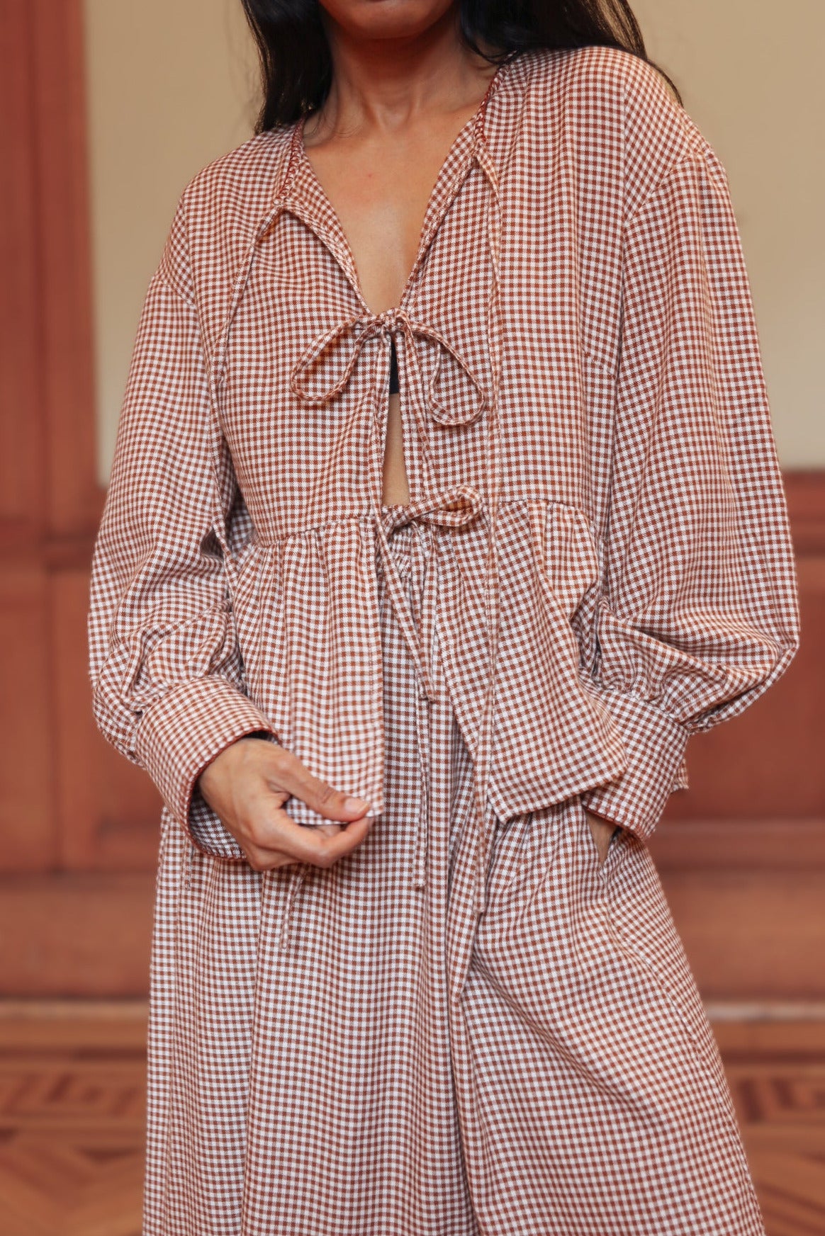 Woman wearing a brown gingham outfit with front tie-ups in a room with wooden paneling.