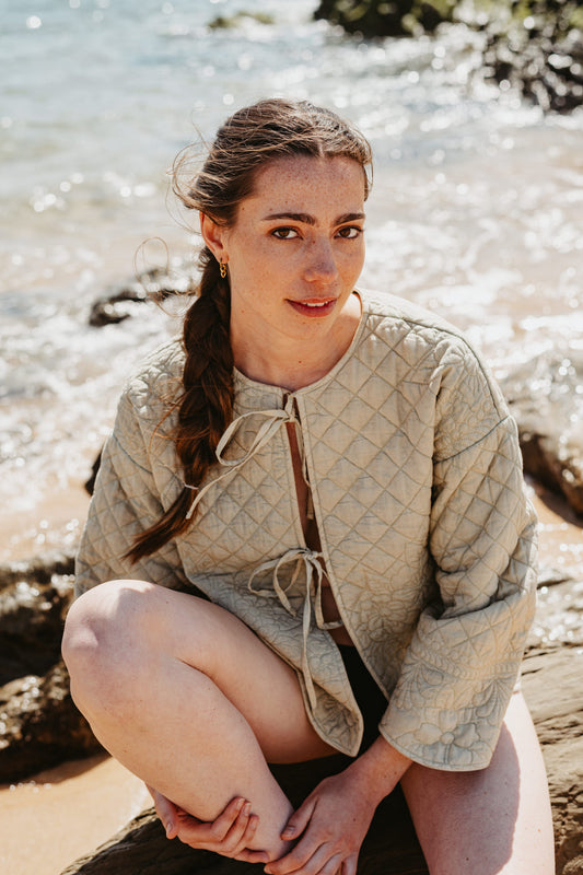 Woman sitting on a log by the sea wearing a green quilted jacket.