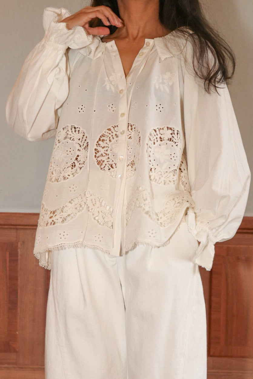 Woman wearing a white blouse with lace details and white pants in a room with wooden paneling.