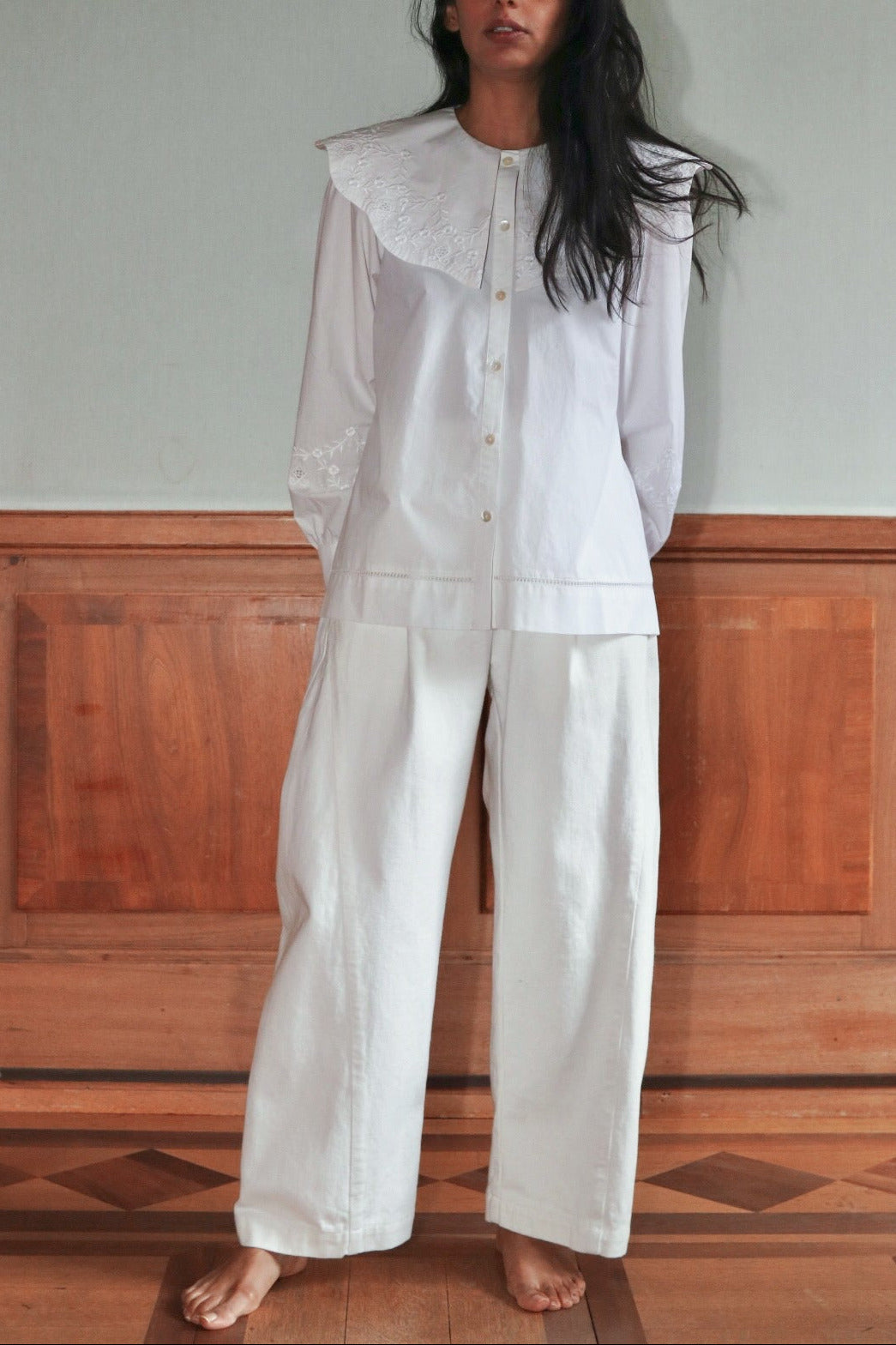 Woman wearing a white shirt and trouser outfit with an embroidered collar against a wooden paneled wall.