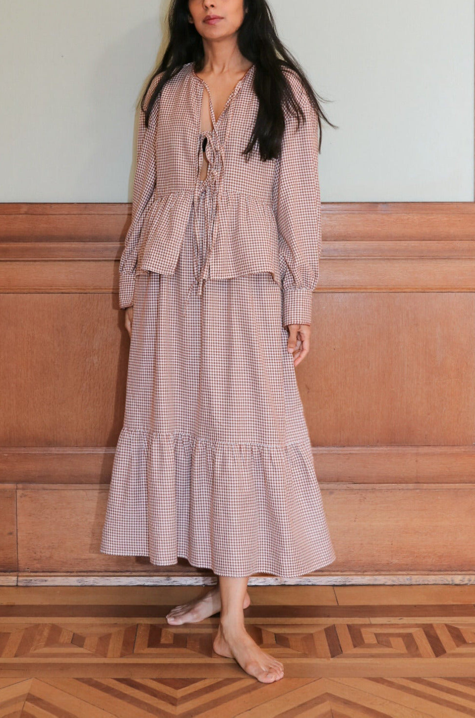 Woman in a brown gingham outfit standing against a wooden paneled wall.