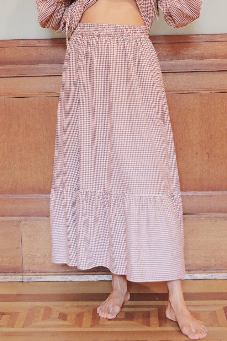 Woman in a brown gingham skirt standing against a wooden paneled wall.