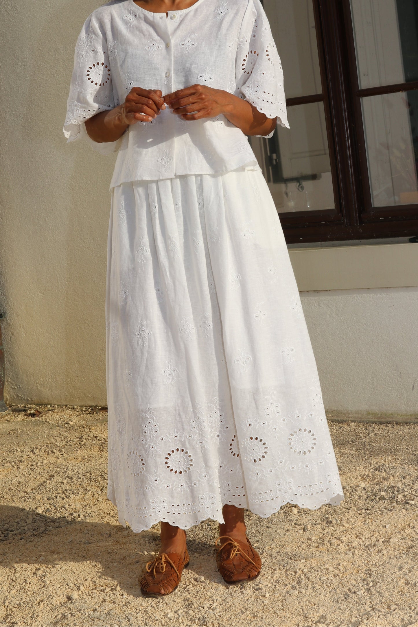 Woman wearing a white dress with intricate patterns standing against a neutral wall.