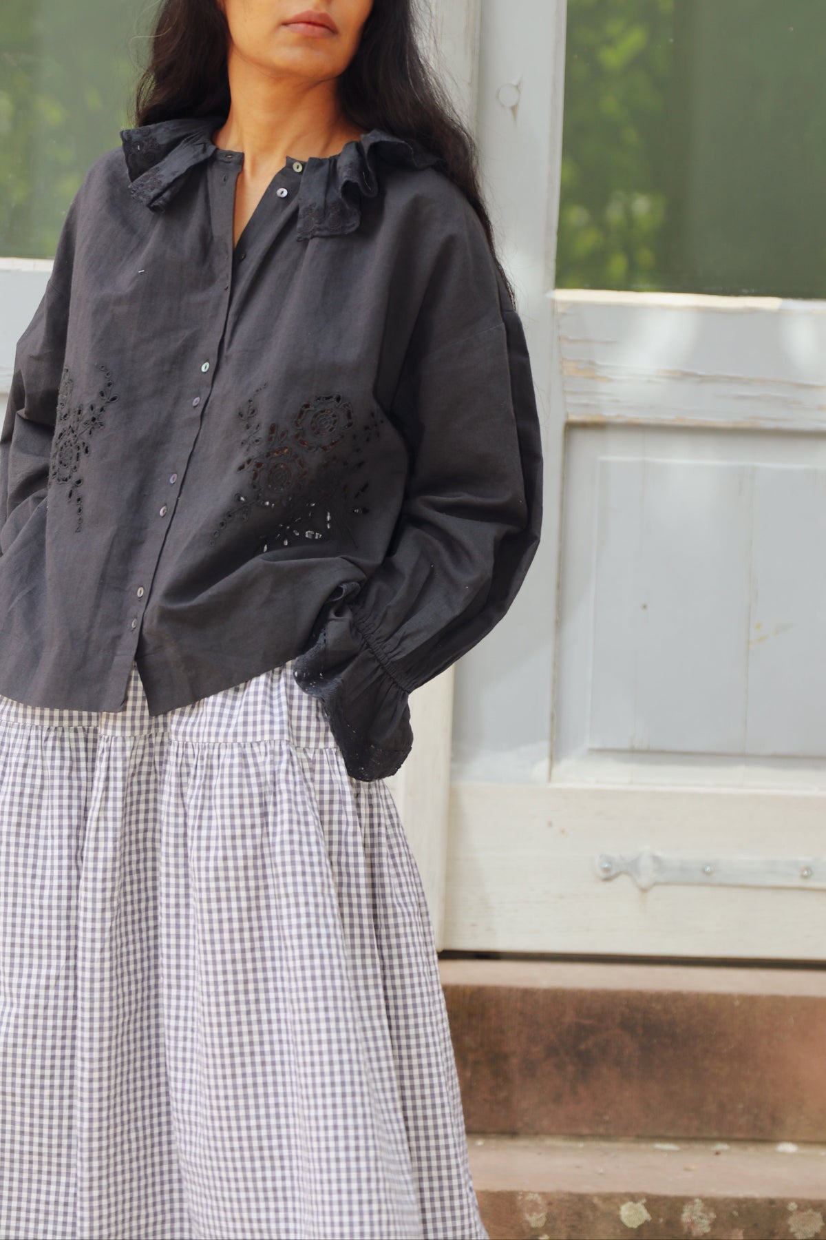 A woman wearing a loose black button-up shirt with embroidered details on the front and a light gray and white gingham skirt, standing in front of a white wooden door.