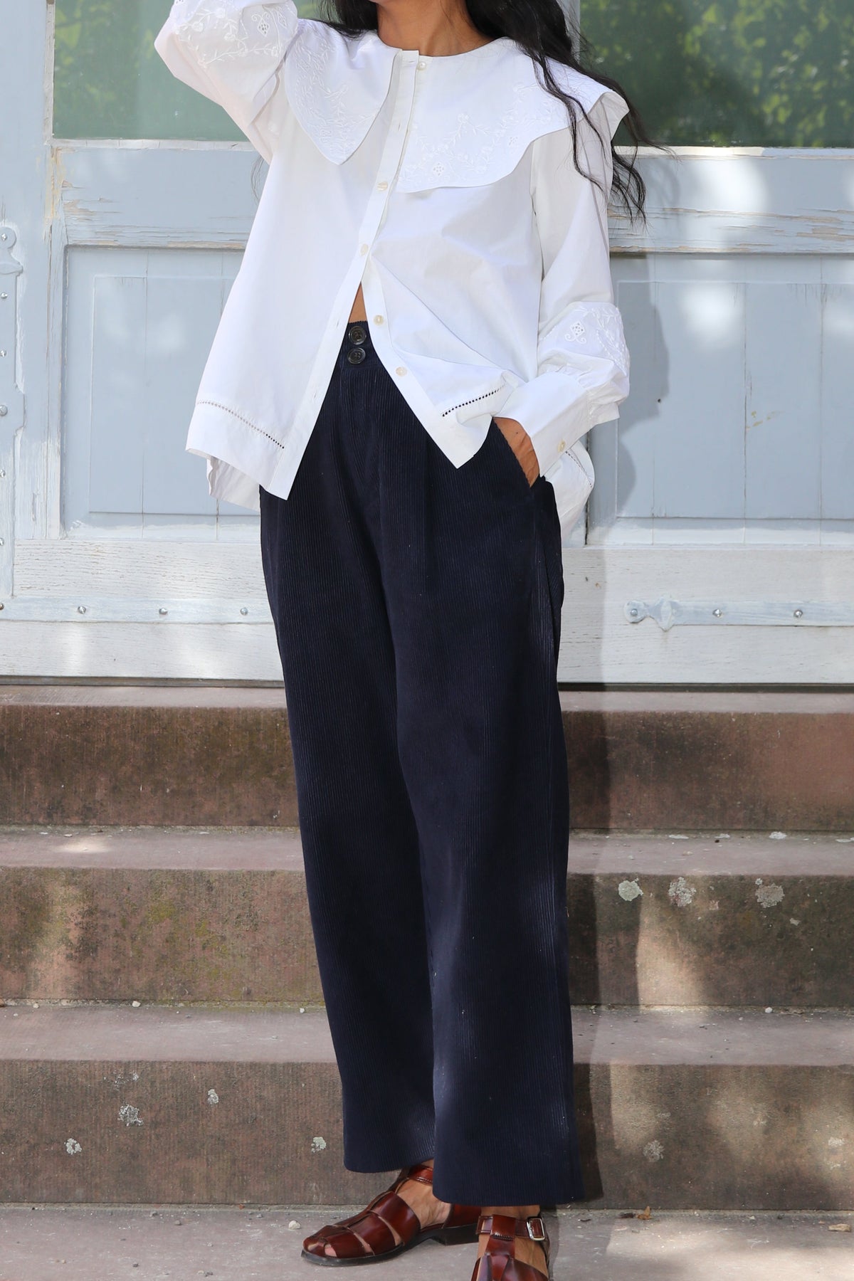 A woman stands on stone steps in front of a light blue door, wearing a white ruffled long-sleeve blouse, dark navy wide-leg trousers, and brown strappy sandals.