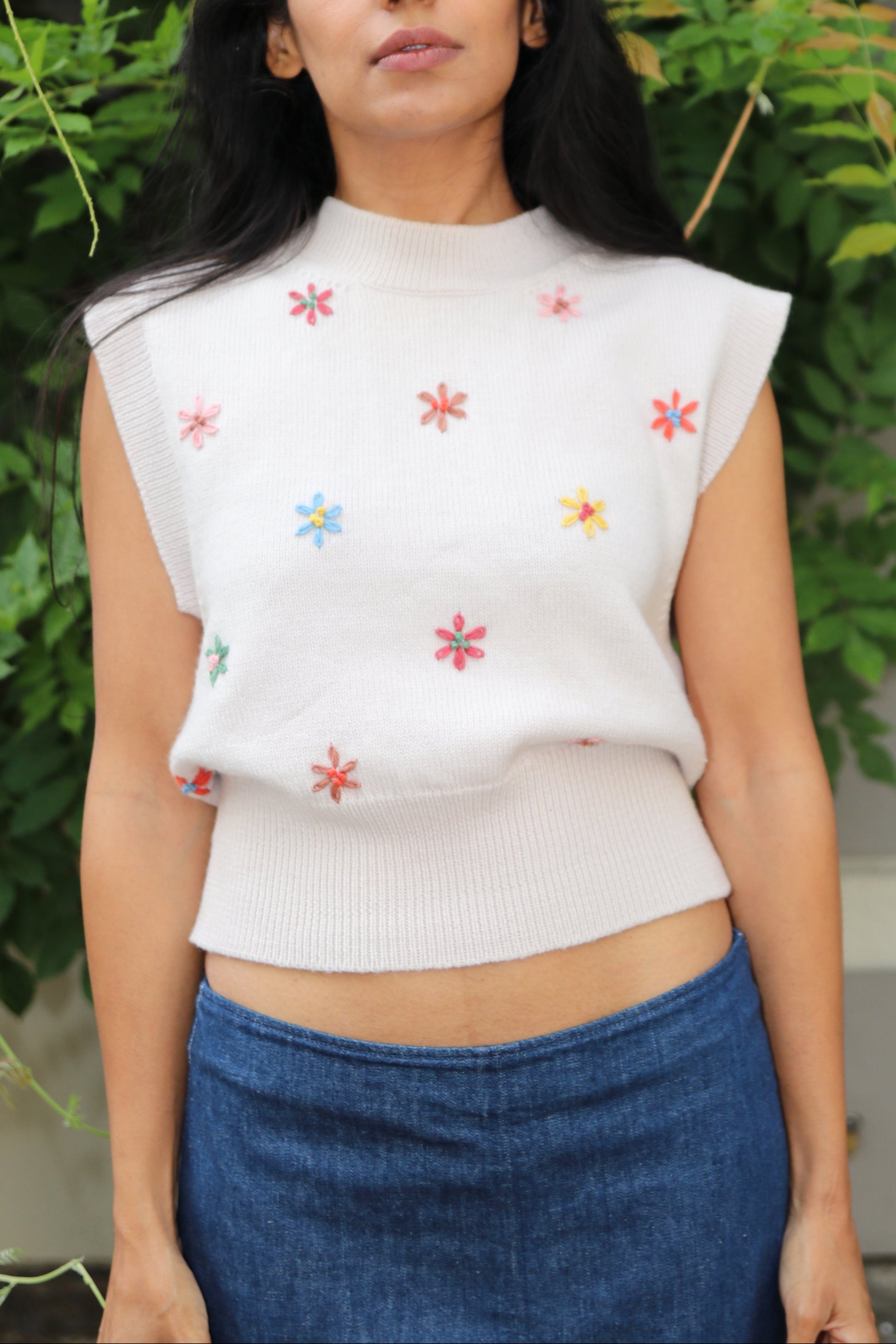 Woman wearing a white top with floral embroidery and blue skirt standing among greenery.