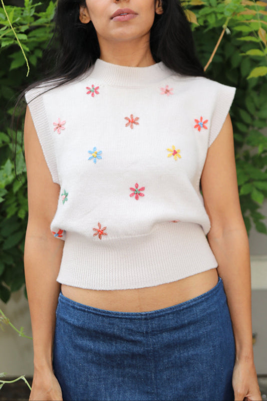 Woman wearing a white top with floral embroidery and blue skirt standing among greenery.