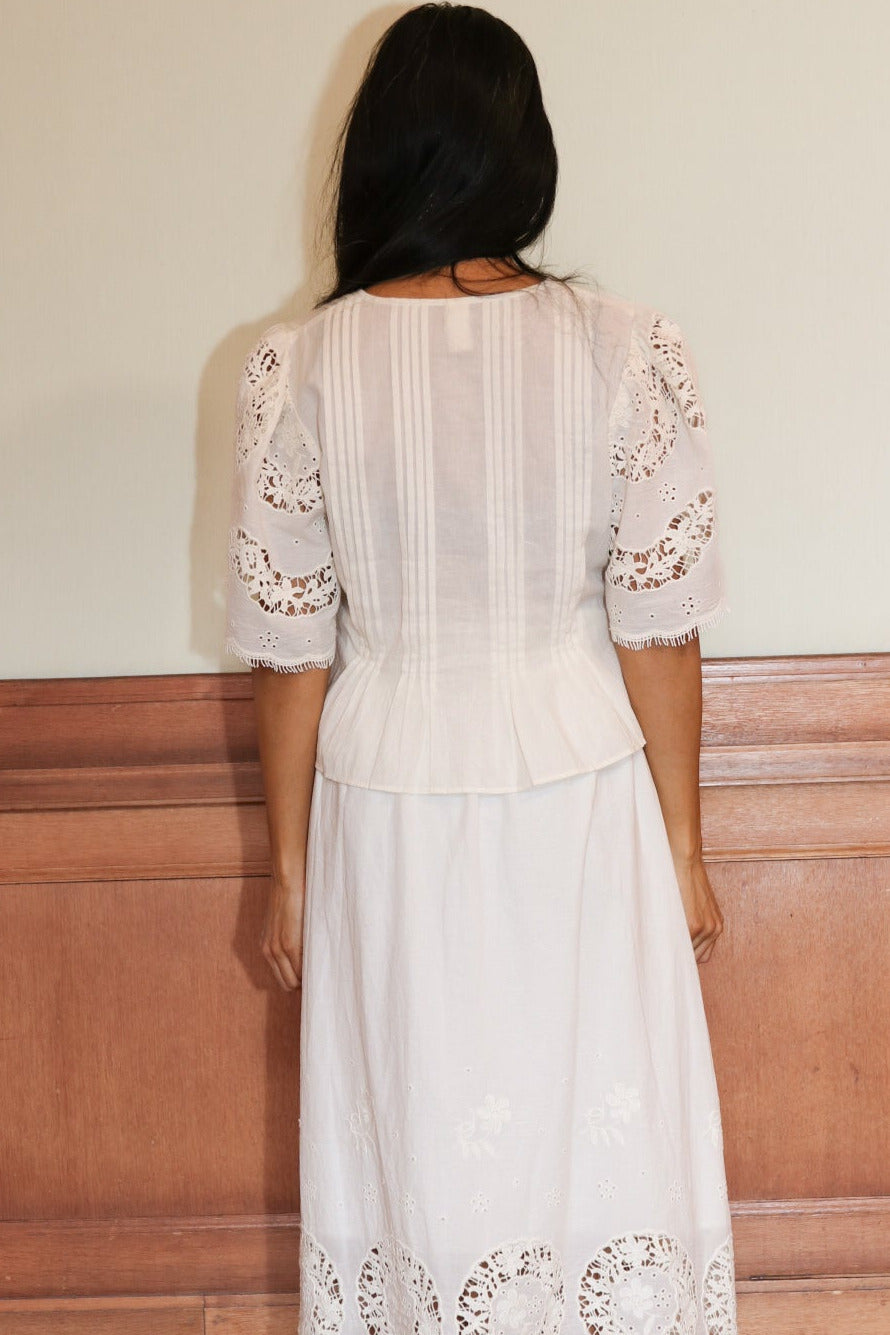 Woman wearing a white lace dress standing against a wooden paneled wall.