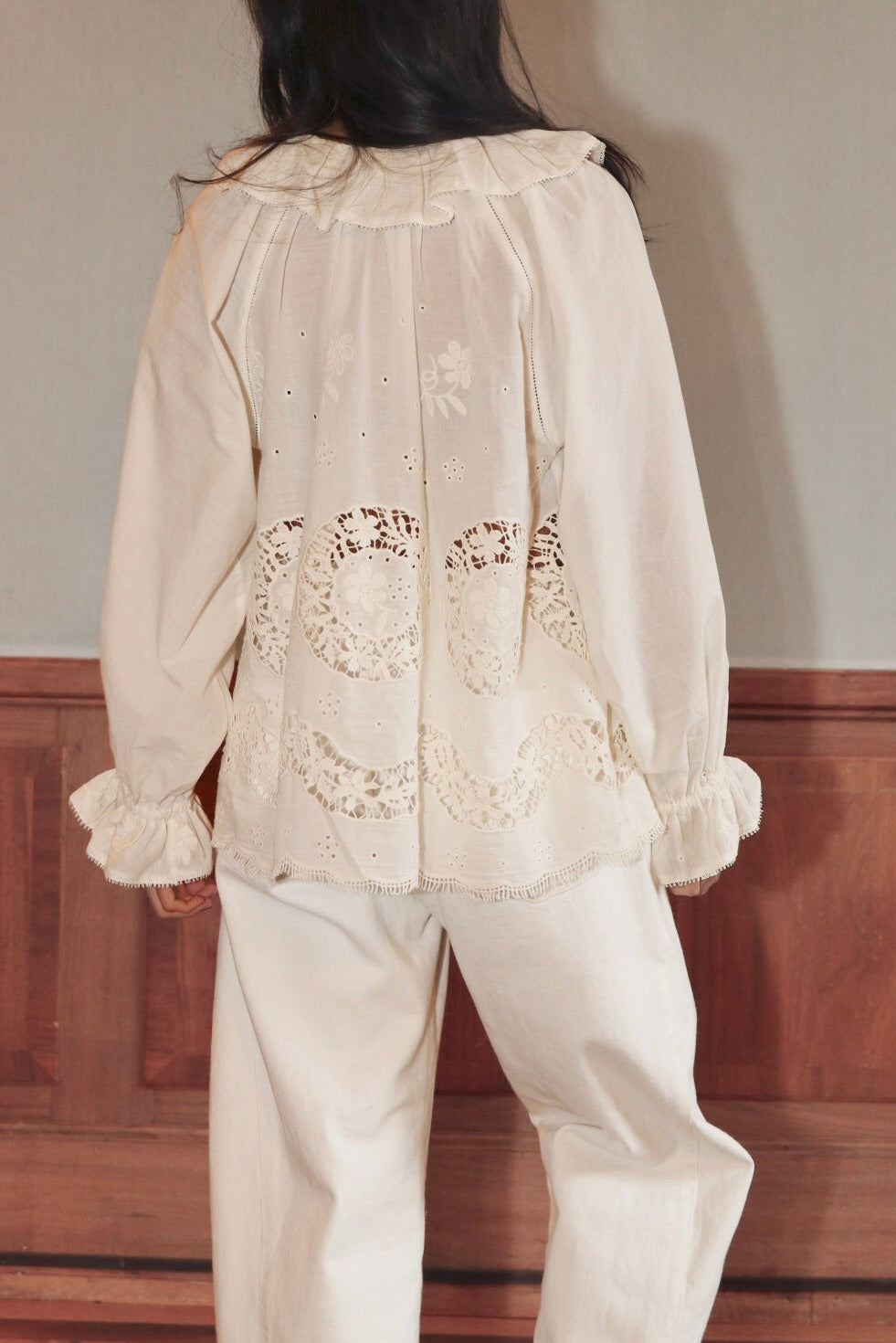 Person wearing a white embroidered blouse and pants against a plain wall.