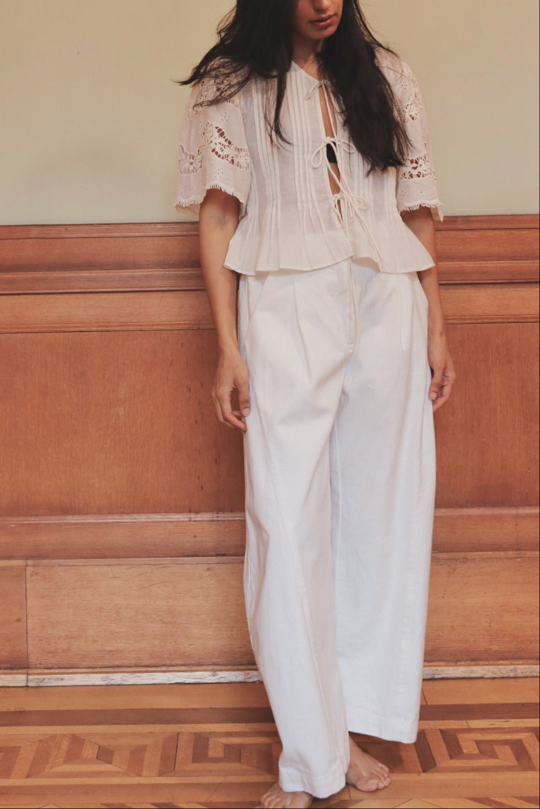 Woman wearing a white outfit with lace details against a wooden paneled wall.