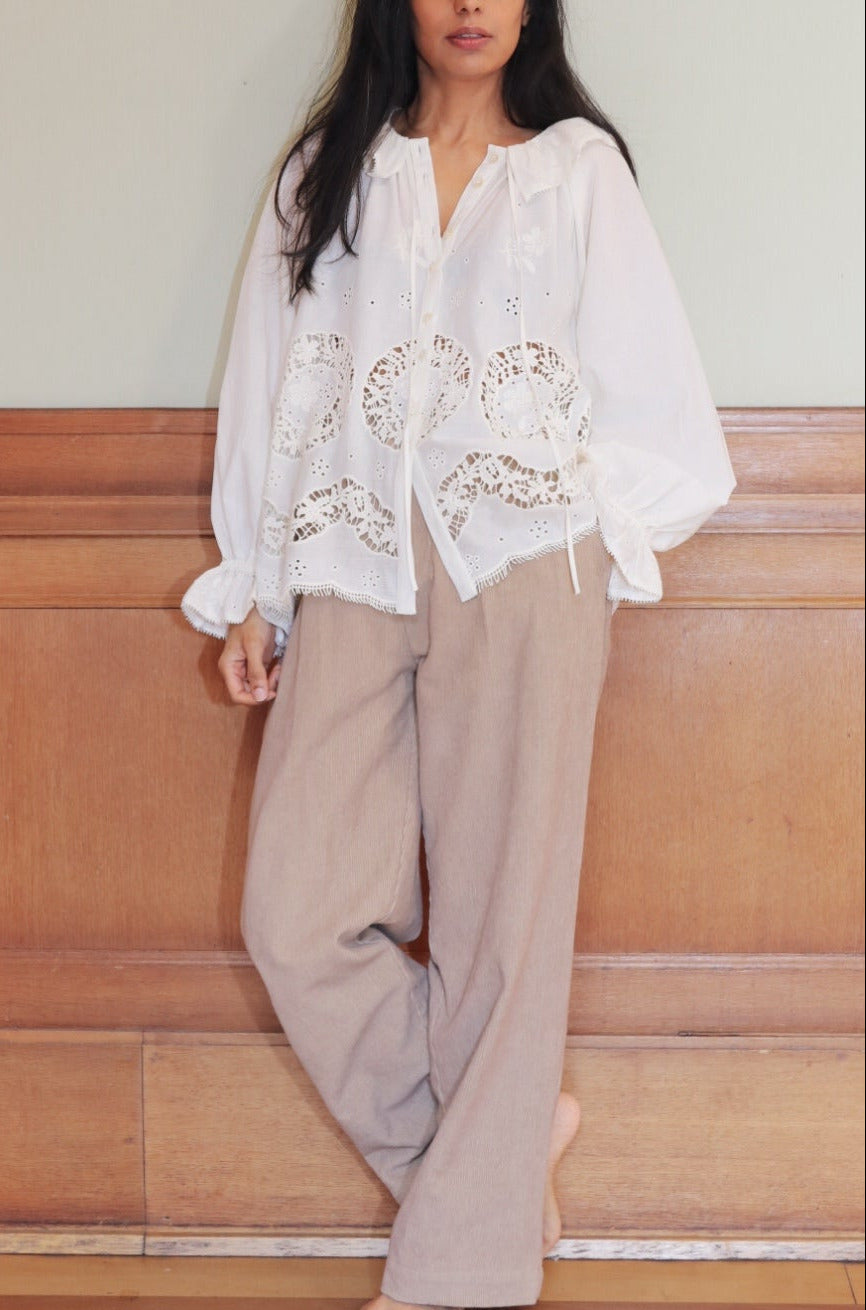 Woman wearing a white blouse with floral embroidery and beige pants against a wooden paneled wall.