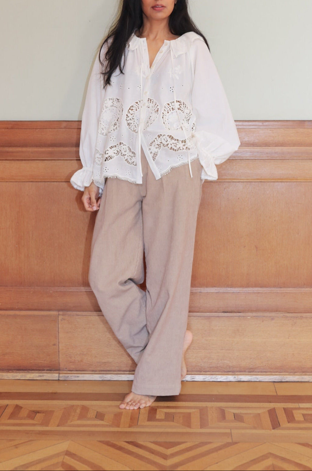 Woman wearing a white blouse with floral embroidery and beige pants against a wooden paneled wall.