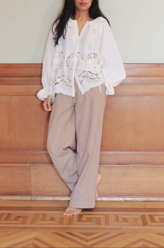 Woman wearing a white blouse with floral embroidery and beige pants against a wooden paneled wall.