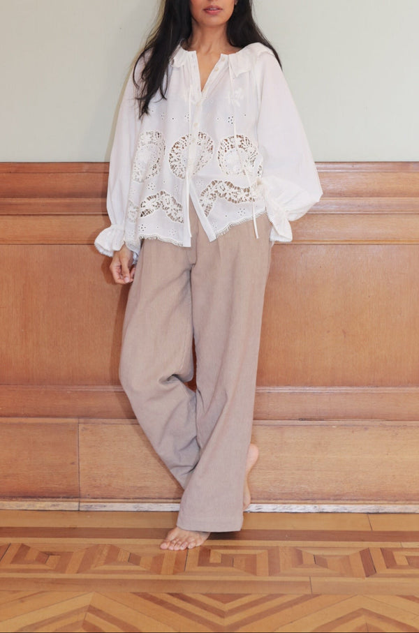 Woman wearing a white blouse with floral embroidery and beige pants against a wooden paneled wall.