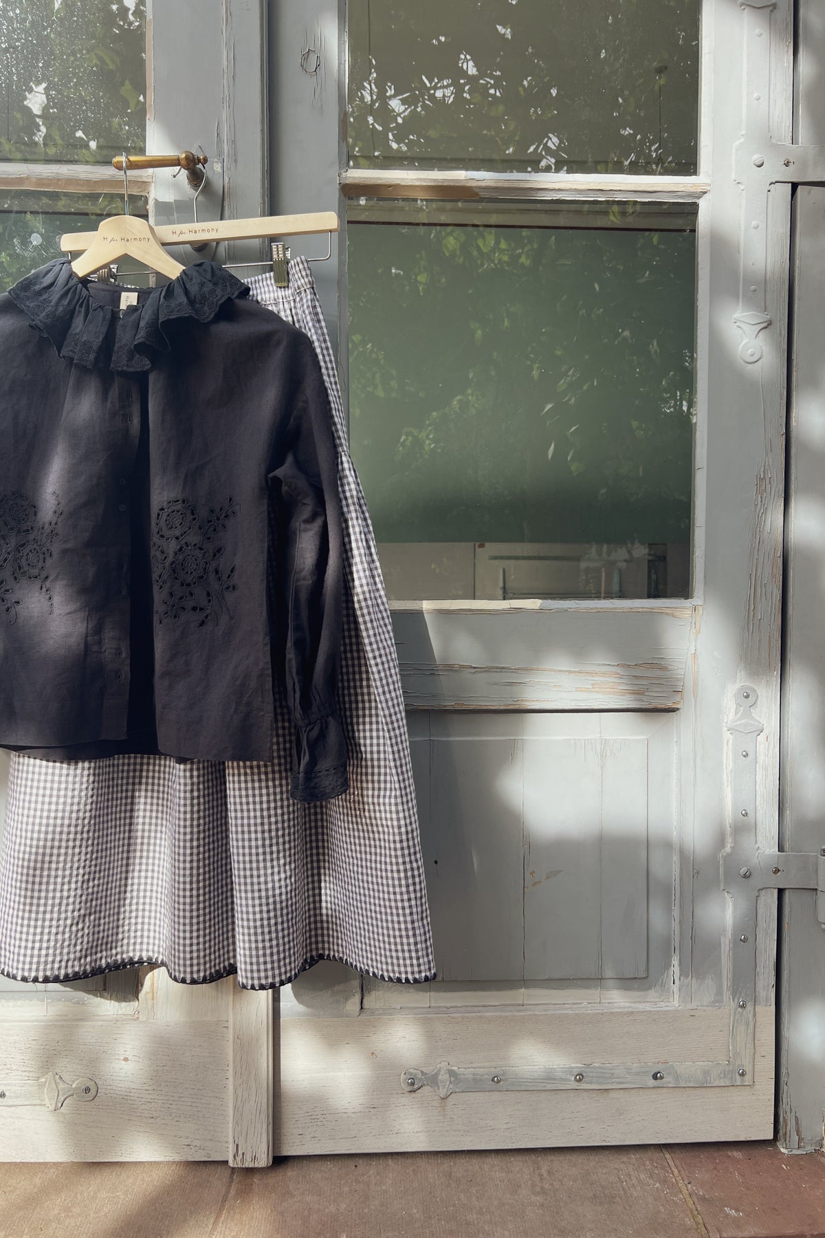 A black embroidered long-sleeve blouse with a ruffled collar hangs on a wooden hanger from a rustic weathered gray door, paired with a black and white gingham skirt.