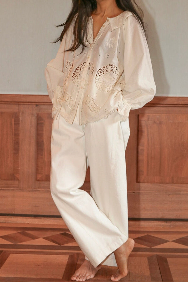 Woman wearing a white embroidered top and pants standing against a wooden paneled wall.