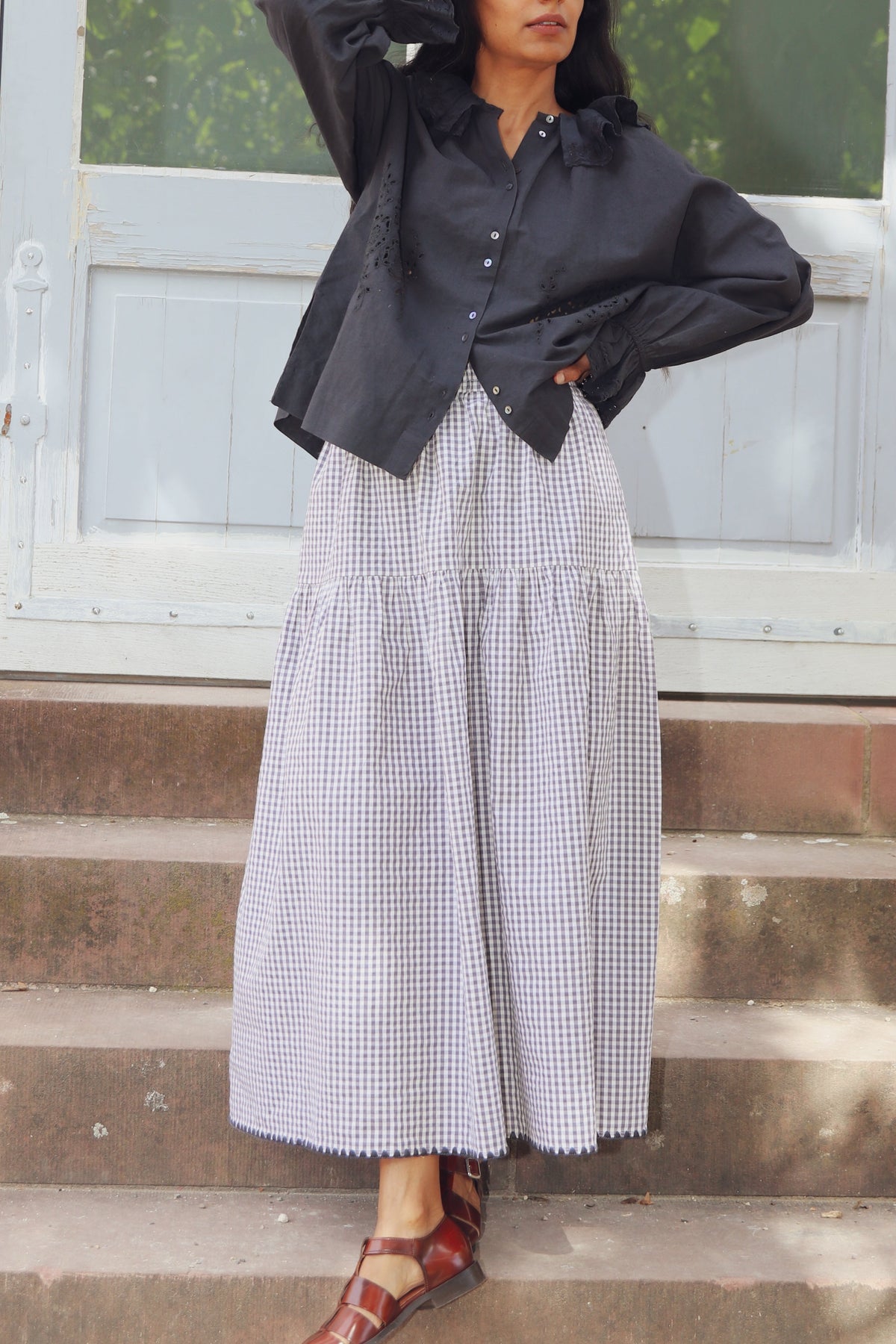 A woman stands on outdoor stone steps in front of a large light blue door, wearing a black long-sleeve button-up shirt and a black-and-white gingham tiered maxi skirt, paired with brown strappy sandals.