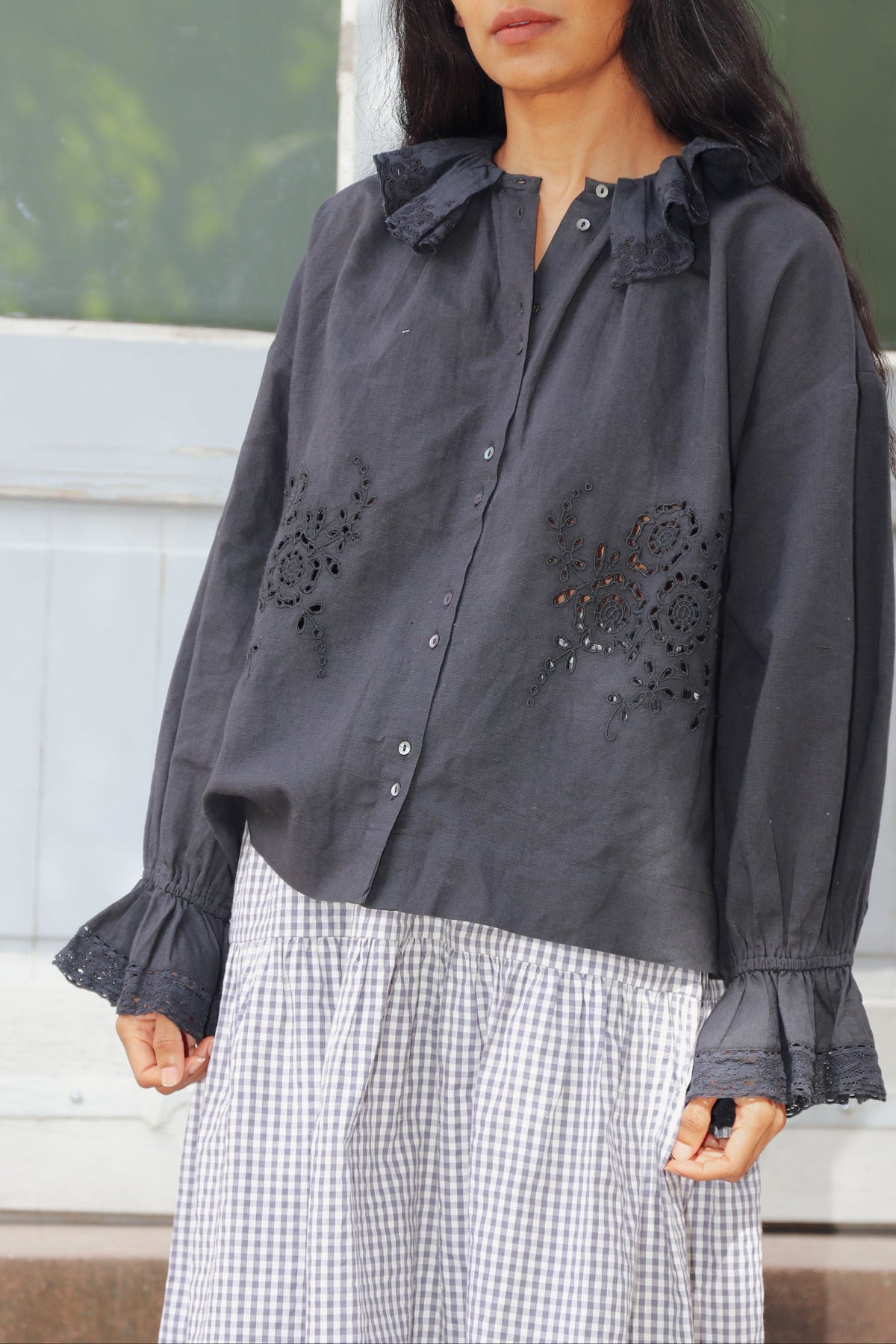 A woman wearing a dark gray long-sleeve button-up blouse with floral embroidery and ruffled collar/cuffs, paired with a white and gray gingham skirt, standing outdoors.